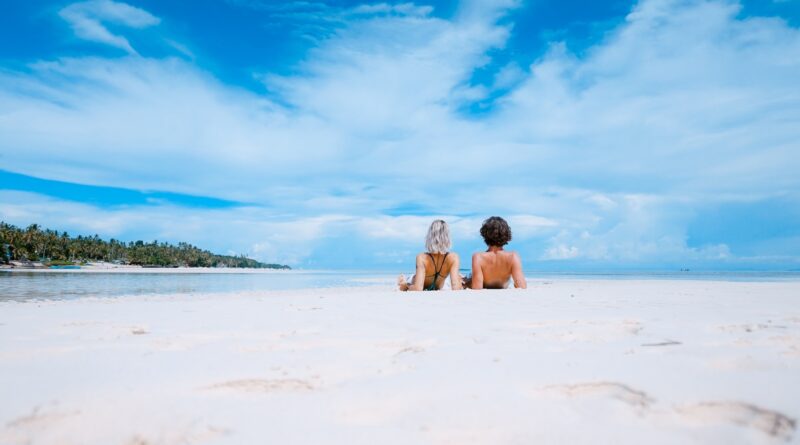 two women lying on white sand facing beach under blue sky